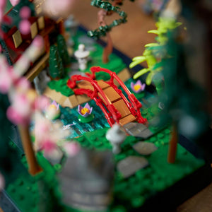 Close-up of a LEGO model with a red bridge, green trees, and small figures on a blurred background.
