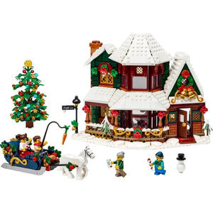 [built-white] A LEGO Christmas village set featuring a snow-covered holiday house with green walls, red wreaths, a decorated Christmas tree, a sleigh with LEGO minifigures, and small accessory figures on a white background.