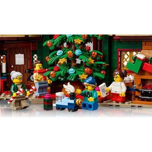 [built-detail] LEGO minifigures arranged around a decorated Christmas tree in a holiday village scene, with gift boxes and a house setting visible in the background.