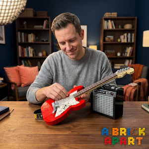 Man playing a red guitar with a small amplifier on a wooden table, with 'A Brick Apart' branding.