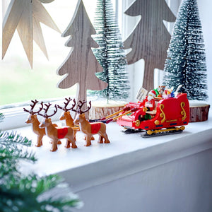 Toy Santa Claus in a sleigh with reindeer on a windowsill with decorative trees in the background.