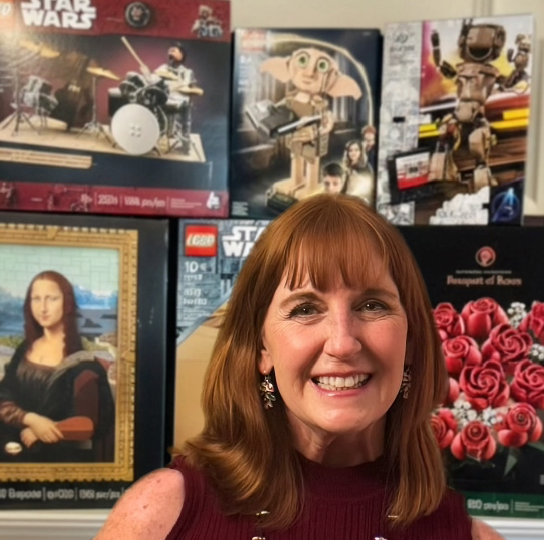 A smiling woman with shoulder-length auburn hair stands in front of a wall displaying various boxed LEGO Star Wars and other toy sets, including a Leonardo da Vinci painting set and a rose set.