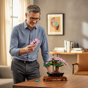 A Brick Apart man assembling a LEGO Bonsai Tree with pink cherry blossoms and green leaves on a wooden desk.