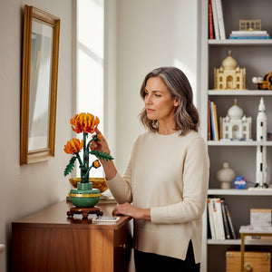 A Brick Apart LEGO Icons Chrysanthemum set with orange flowers in a green pot, displayed by a woman in a home interior.