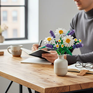 A Brick Apart LEGO Botanicals Daisies bouquet with lavender in a white vase on a desk with a person writing nearby.