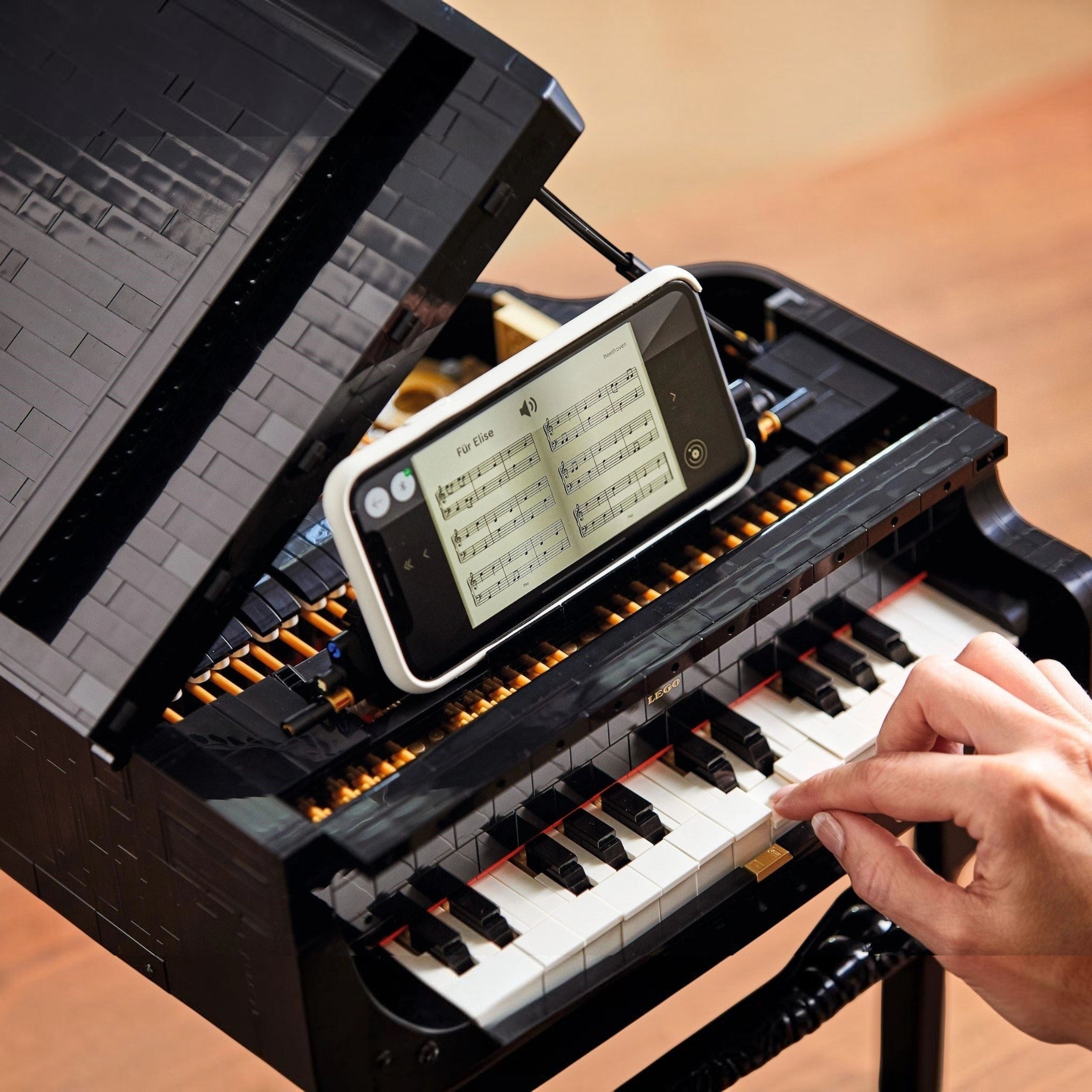 A Brick Apart LEGO Ideas Grand Piano set with a smartphone displaying sheet music on the stand, being played by hand.