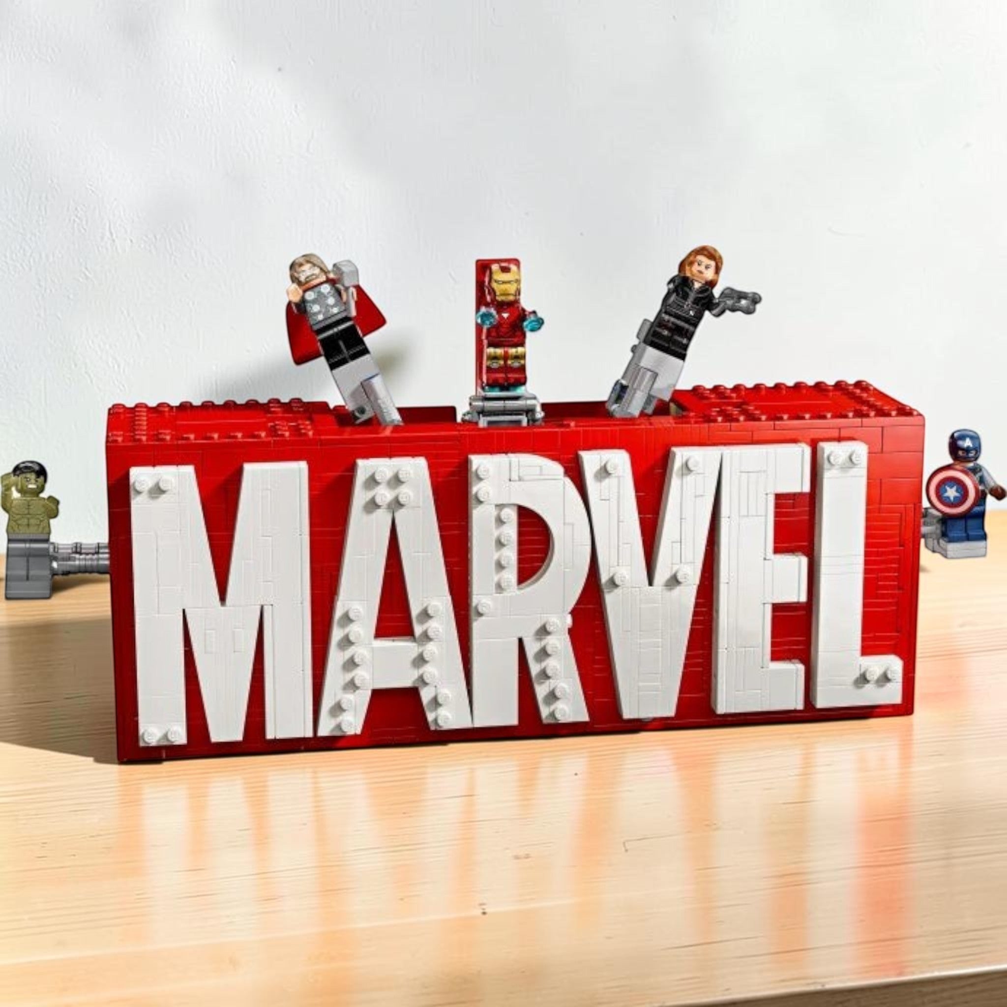 A Brick Apart LEGO Marvel logo display set featuring red and white bricks with Hulk, Thor, Iron Man, Black Widow, and Captain America minifigures.
