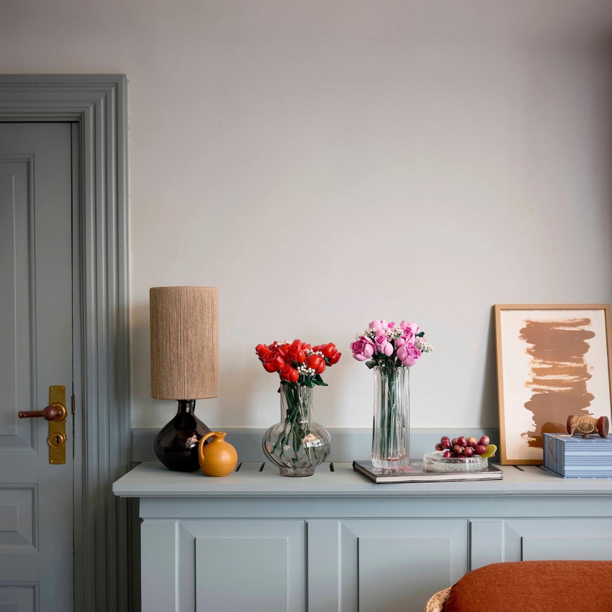 A Brick Apart LEGO Botanicals Bouquet of Pink Roses displayed in a glass vase on a blue console table with home decor.
