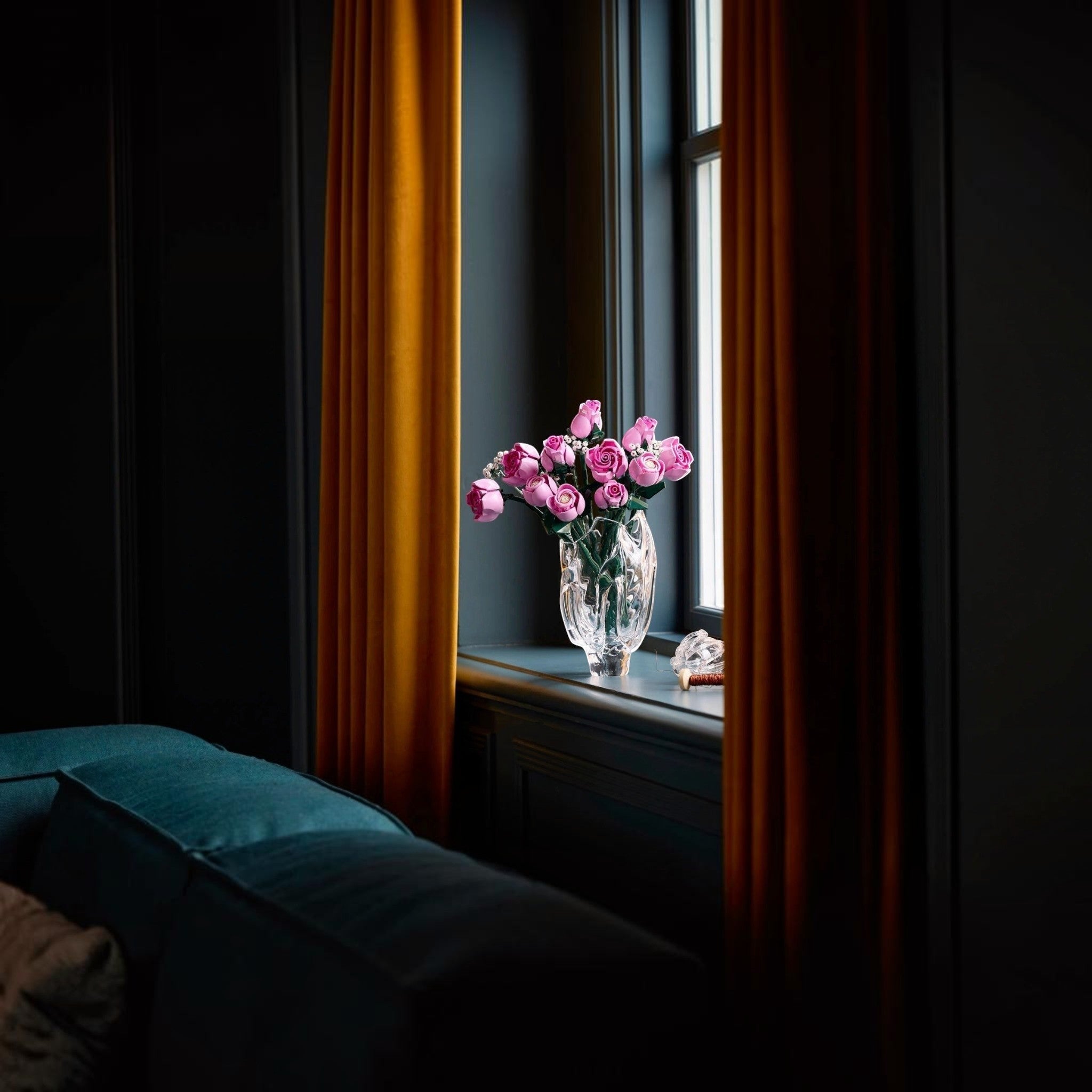 A Brick Apart LEGO Botanicals bouquet of pink roses in a glass vase displayed on a windowsill with mustard curtains.
