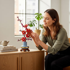 A Brick Apart LEGO Icons Plum Blossom set with red flowers in a blue pot, displayed on a wooden table by a woman.