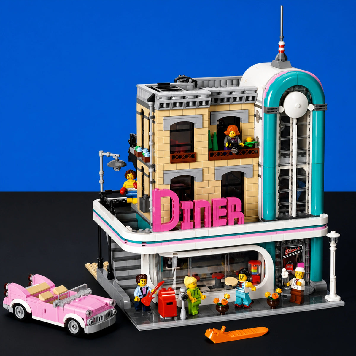 [built-blue-black] A detailed toy model of a retro-style diner building set against a split blue and black background. The set includes a beige and gray multi-story building with a prominent pink 'DINER' sign, a teal arched tower, a pink convertible car, and several small minifigures and accessories.
