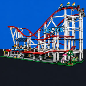 [built-blue-black] A large assembled roller coaster toy structure made of interlocking building blocks, shown against a blue-to-black gradient background. The coaster has red tracks, a white support frame, green base details, and small multicolored minifigures and ride accessories.