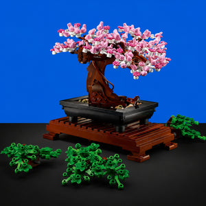 [built-blue-black] A building-block toy bonsai tree with pink and white blossoms, a brown trunk and pot, and green foliage pieces arranged around it on a black surface against a bright blue background.
