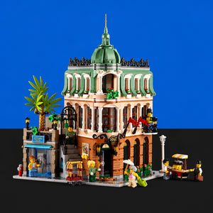 [built-blue-black] A detailed LEGO city building set featuring a multi-story colonial-style building with a green dome, beige and brown facade, a palm tree, street market stalls, and small minifigures, shown against a split blue and black background.