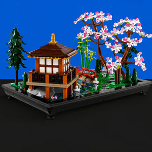 [built-blue-black] A detailed building-block set depicting a Japanese garden scene on a rectangular base. The model includes a brown pagoda-style pavilion, a red arched bridge, a flowering cherry tree with pink and white blossoms, green bamboo, small stone lanterns, and a pond with pink lotus flowers.