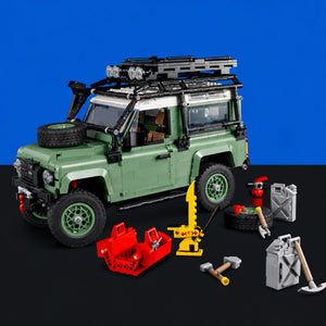 [built-blue-black] A green brick-built off-road vehicle set with a black roof rack, spare tire, and accessories arranged on a black surface against a blue background. The set includes red tools, a yellow crane, gray crates, and small toy vehicles.