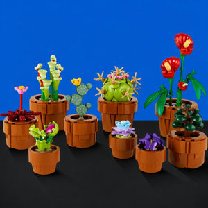 [built-blue-black] A set of small building-block plant toys in terracotta-colored pots, arranged on a black surface against a bright blue background. The collection includes cacti, succulents, flowers, and leafy stems in green, red, purple, and pink.