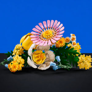 [built-blue-black] A colorful floral arrangement of artificial flowers and leaves on a black surface against a solid blue background. The arrangement features a large pink daisy-like flower, yellow marigold-style blooms, cream blossoms, green foliage, and small light blue buds.