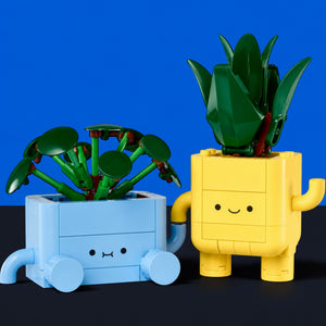 [built-blue-black] Two small block-built character planters on a dark surface against a bright blue background. One planter is light blue and the other is yellow, each holding a green leafy plant.