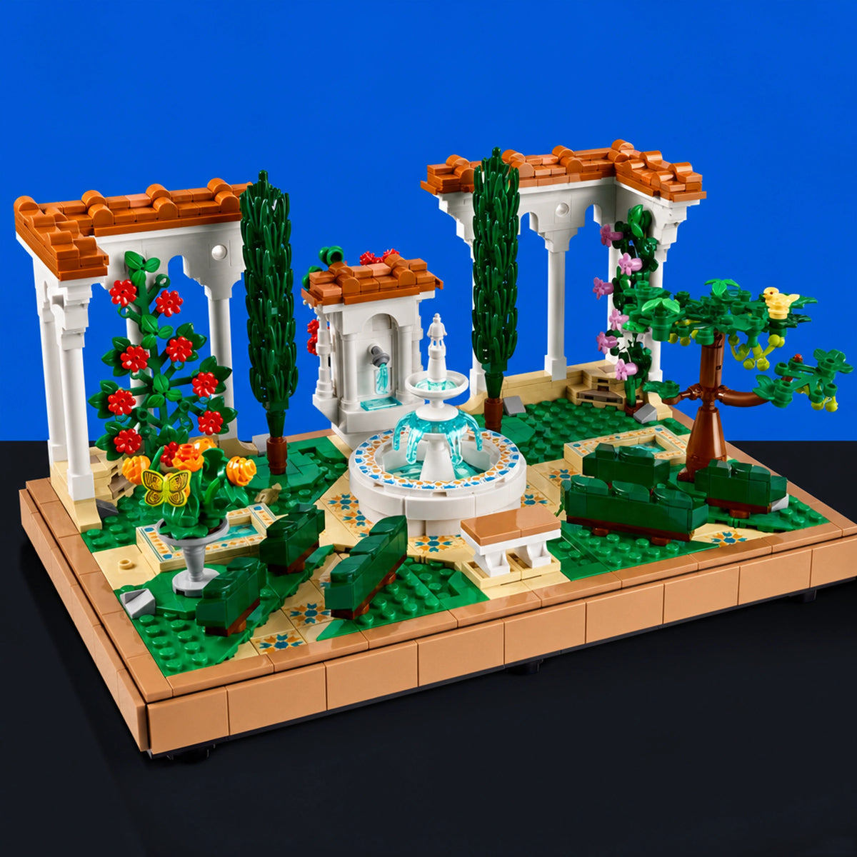 [built-blue-black] A LEGO-style garden and fountain building set displayed on a tan baseplate against a blue studio background. The set includes white arches, a central fountain with blue and white details, green lawn and bushes, trees, flowers, benches, and small architectural elements.