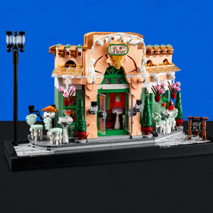 [built-blue-black] A detailed LEGO-style cafe building set displayed on a black base against a blue studio background. The structure is peach and green with white decorative trim, outdoor seating, small trees, and a black street lamp.