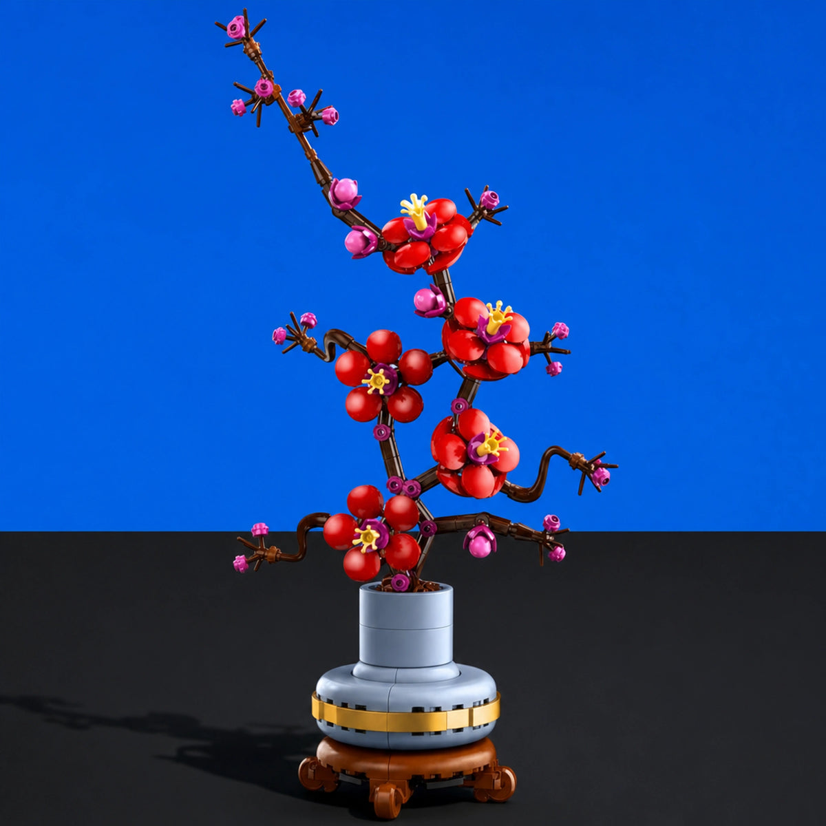 [built-blue-black] A building-block style decorative plum blossom tree sculpture with red blossoms, pink buds, brown branches, a gray cylindrical pot, and a small wooden base, photographed against a blue and black studio background.