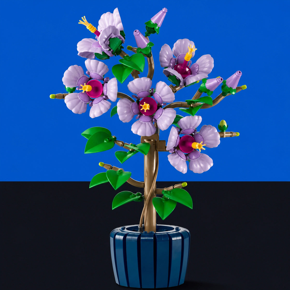 [built-blue-black] A toy building-block style flowering tree in a blue striped pot, shown against a split blue and black background. The flowers are light purple with pink centers and green leaves, mounted on a brown trunk.