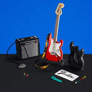 [built-blue-black] A miniature red electric guitar set with a black amplifier, black headstock, black body parts, colorful studs, a turquoise pick, and a Fender sticker on a black surface against a bright blue background.