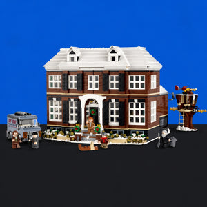 [built-blue-black] A detailed brick-built model of a two-story house with a white roof, brown brick walls, black shutters, and a snowy front porch. A gray tow truck labeled 'TOW JAY' is parked on the left, and a small treehouse structure with a ladder is on the right, all set against a solid blue background with a black floor.