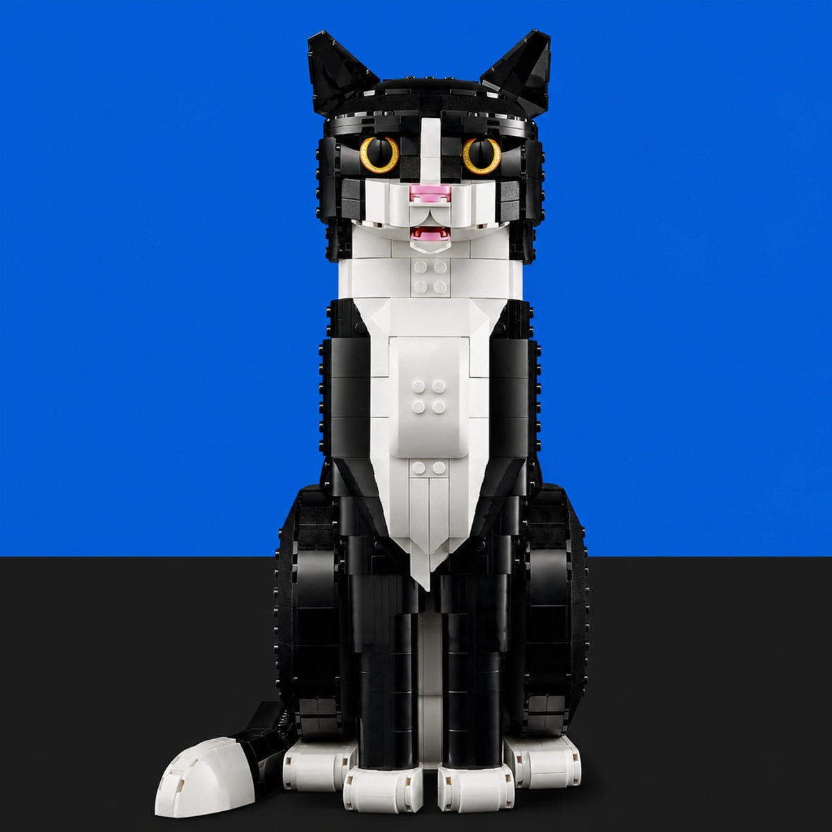 [built-blue-black] A black and white cat-shaped building block toy with yellow eyes, pink nose, and white paws, shown against a split blue and black background.