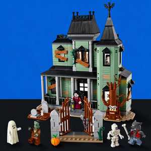 [built-blue-black] A LEGO-style haunted house building set displayed on a black surface against a bright blue background. The set includes a mint green and gray two-story haunted house with dark roof, a gate, spooky tree, pumpkin, and several small minifigures including a ghost, zombie, mummy, catwoman, and vampire.