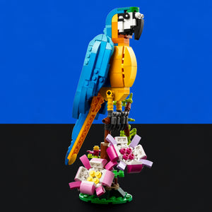 [built-blue-black] A toy macaw bird built from colorful building blocks, perched on a branch with flowers at the base, photographed against a split blue and black background.