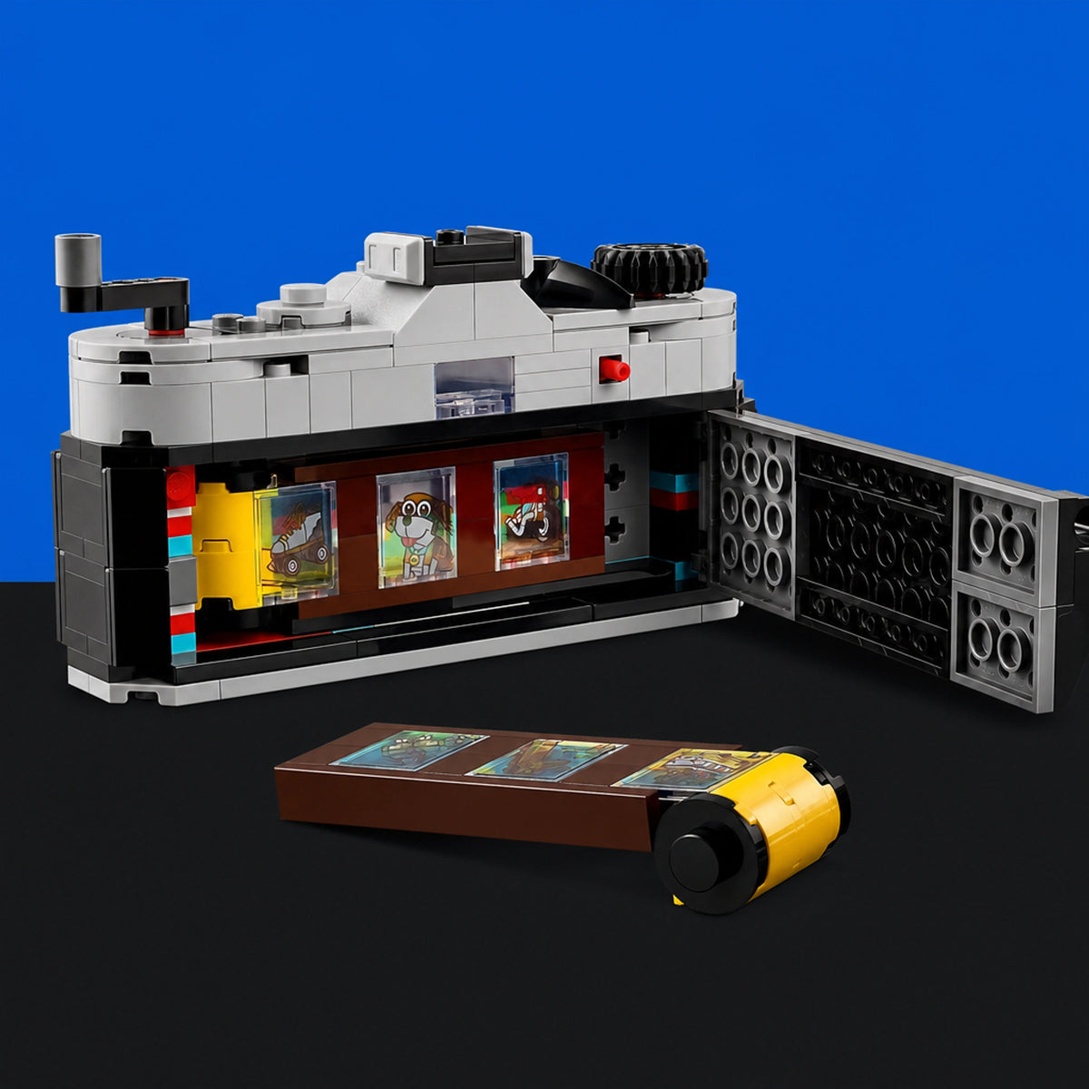 [built-blue-black] A LEGO-style model set displayed on a black surface against a bright blue background, featuring a gray, black, and white structure with colorful panel details and a separate brown-and-yellow accessory piece placed in front.