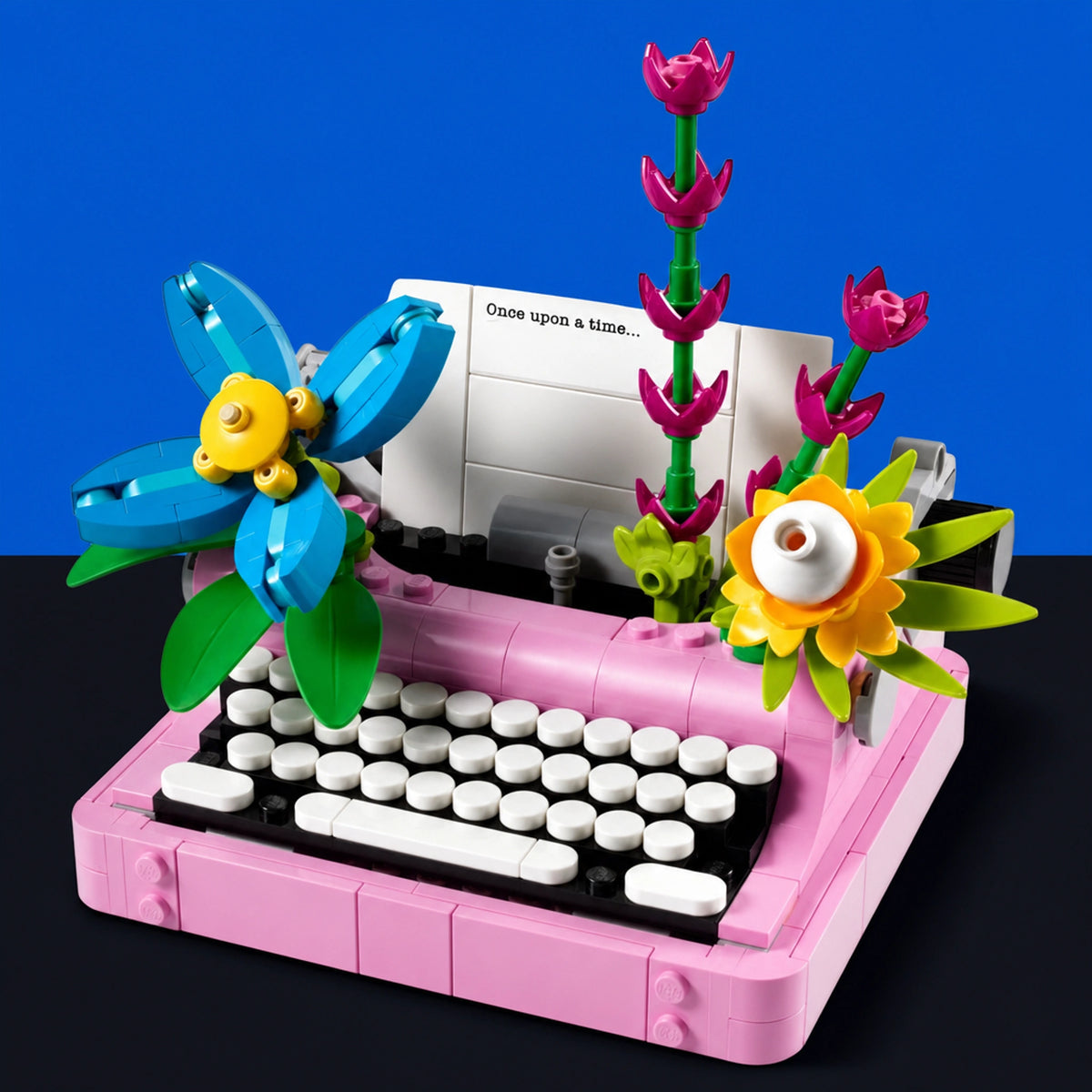 [built-blue-black] A pink toy typewriter built from interlocking blocks, with white keys, a black keyboard base, and decorative flowers growing out of the top. The typewriter is photographed on a dark surface against a bright blue background.