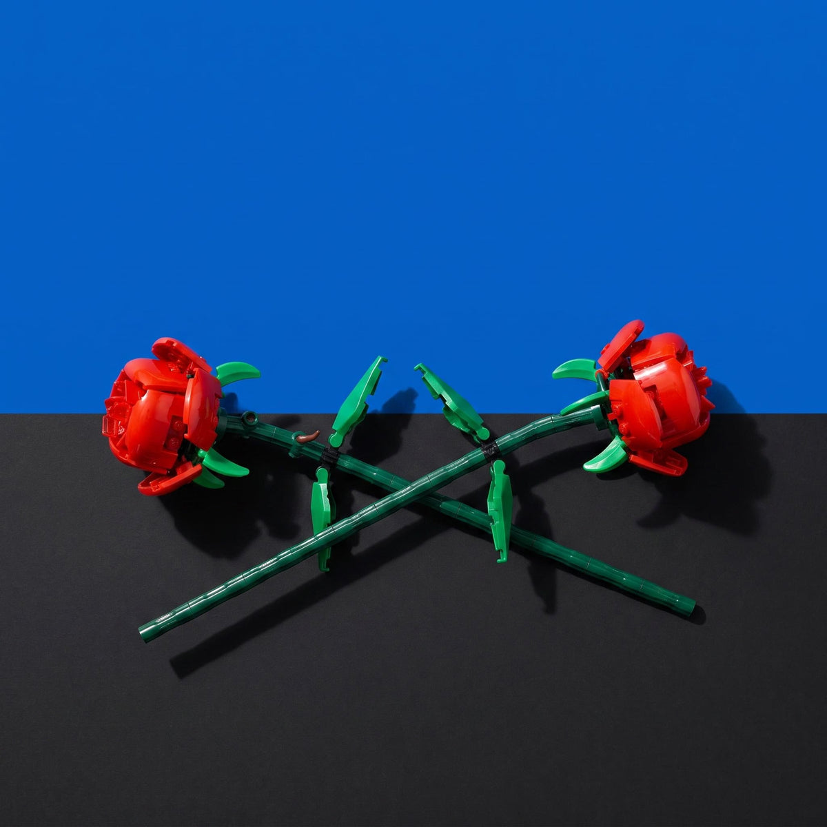 [built-blue-black] Two red rose-shaped items with green stems and leaves, arranged crossed on a black surface against a blue background.
