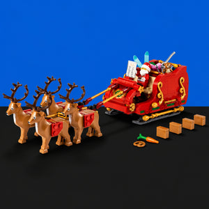 [built-blue-black] A red Santa Claus sleigh toy with four brown reindeer, shown on a black surface against a bright blue background. The set includes small accessory pieces and boxes placed in front of the sleigh.