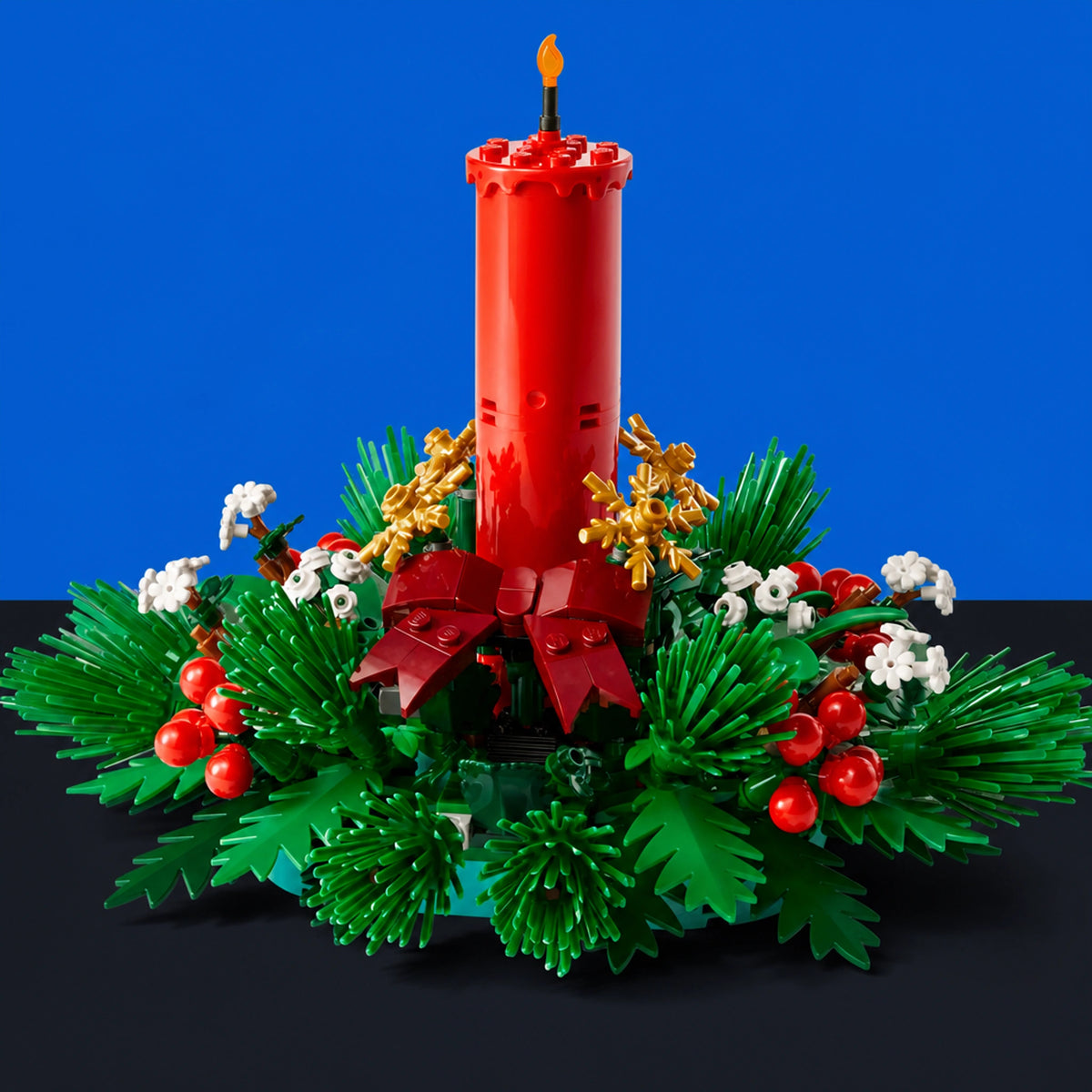 [built-blue-black] A Christmas Advent wreath centerpiece built from LEGO, featuring a tall red pillar candle with a small yellow flame, green pine-like foliage, red berries, white flowers, and gold ornaments, set against a solid blue background.