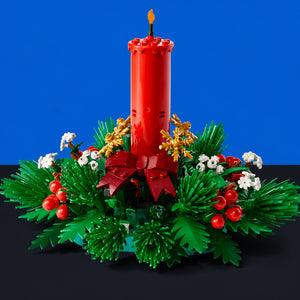 [built-blue-black] A Christmas Advent wreath centerpiece built from LEGO, featuring a tall red pillar candle with a small yellow flame, green pine-like foliage, red berries, white flowers, and gold ornaments, set against a solid blue background.