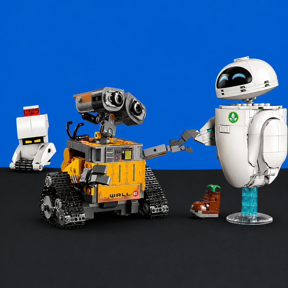 [built-blue-black] A toy building-block set featuring WALL-E and EVE, with WALL-E in yellow and gray on a black surface against a blue backdrop, plus small accessory pieces including a boot and a small white figure.