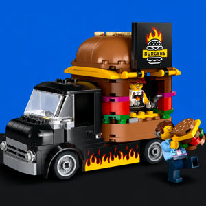 [built-blue-black] A toy construction-style food truck set with a burger-themed cargo container, flame decals, a black cab, and a small minifigure standing beside it holding a burger, photographed against a solid blue studio background.