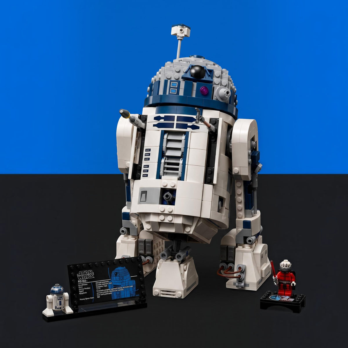 [built-blue-black] A LEGO Star Wars R2-D2 set displayed on a black surface against a blue and black background. The set includes the assembled white, blue, and gray droid, a small R2-D2 figure, a red-and-white minifigure, and a black display card.