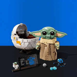 [built-blue-black] LEGO Star Wars The Child (Grogu) set displayed with its assembled green-and-tan toy figure, a white egg-shaped vehicle, a small blue rover, a display plaque, and a gray sphere on a black surface against a blue studio background.
