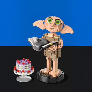 [built-blue-black] A toy figure resembling Dobby the Dragon from Harry Potter, built from interlocking blocks, stands on a black display base next to a small decorated cake on a clear stand. The background is split horizontally with a bright blue upper half and a black lower half.