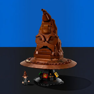 [built-blue-black] A LEGO Harry Potter Hogwarts crest set displayed on a black surface against a blue background, featuring a brown pointed hat structure, a round brown base, a small minifigure, and a small yellow crest piece.