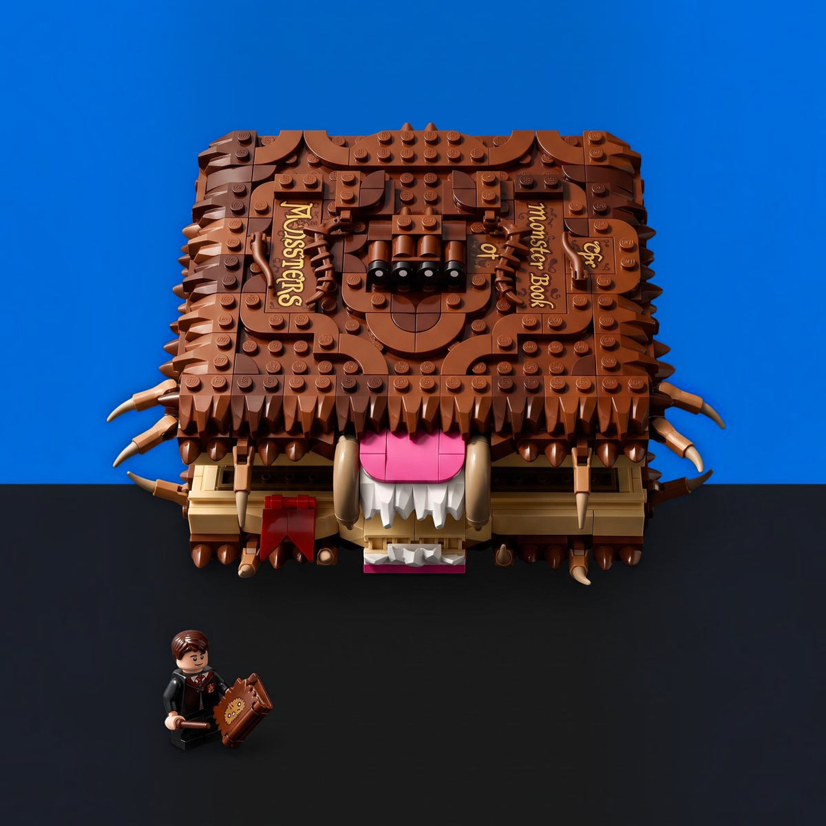 [built-blue-black] A LEGO-style monster book toy with a brown textured cover, gold lettering, a pink mouth, and small horns, shown on a black surface against a blue background. A small minifigure sits at the bottom left holding a book.