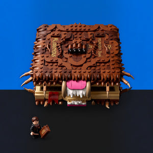 [built-blue-black] A LEGO-style monster book toy with a brown textured cover, gold lettering, a pink mouth, and small horns, shown on a black surface against a blue background. A small minifigure sits at the bottom left holding a book.